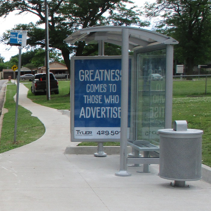A bus shelter is seen from the right side with a trash receptacle on the right, bench seating inside the shelter and a left side panel advertising display.