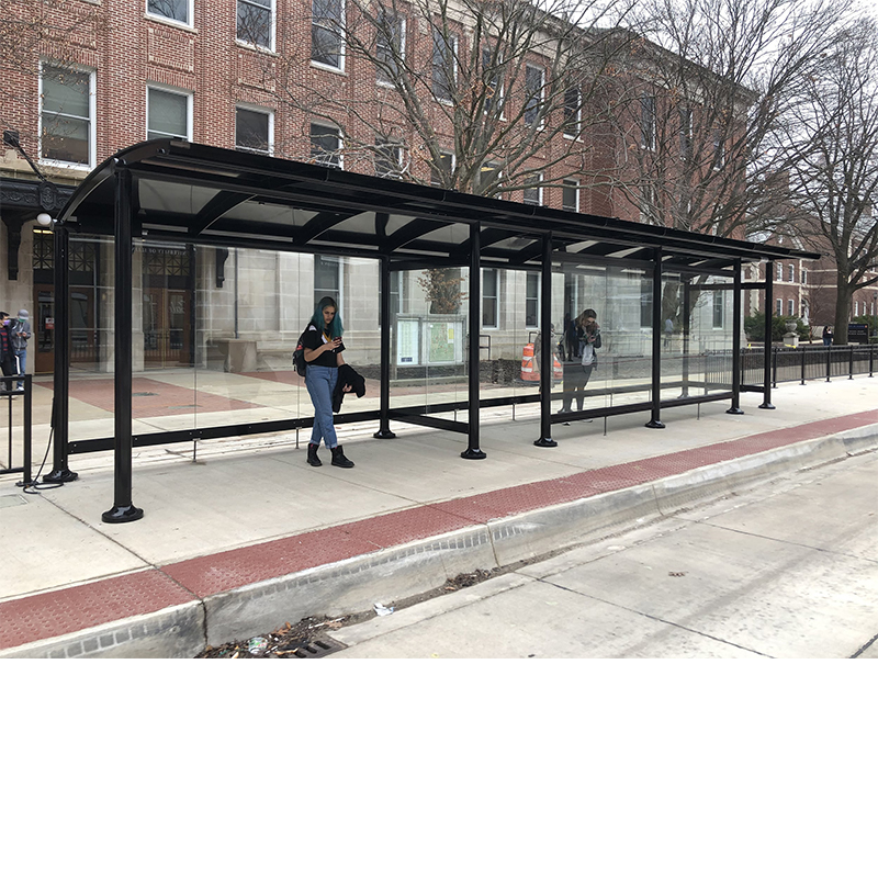 A bus shelter with two riders waiting on each side of the shelter with one side being fully enclosed and one is open to the elements.