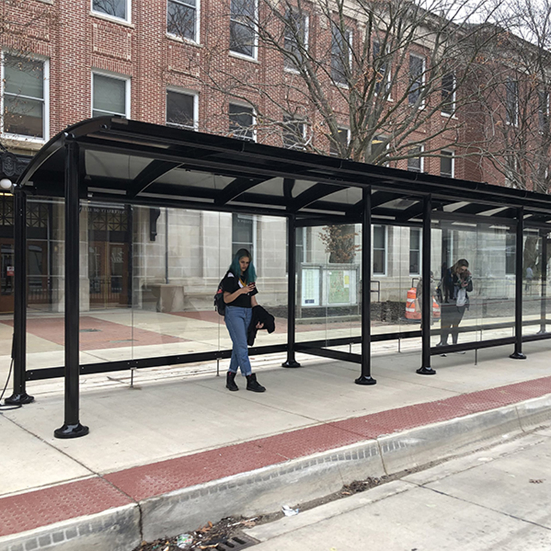 A signa sunset bus shelter with glass siding where two riders await the bus with one side having a one panel wide opening to shelter riders from the elements.
