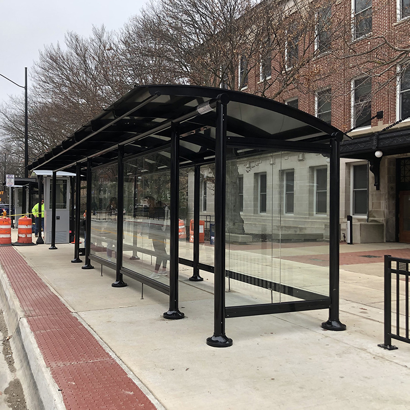 A bus shelter with two riders waiting on each side of the shelter with one side being fully enclosed and one is open to the elements.
