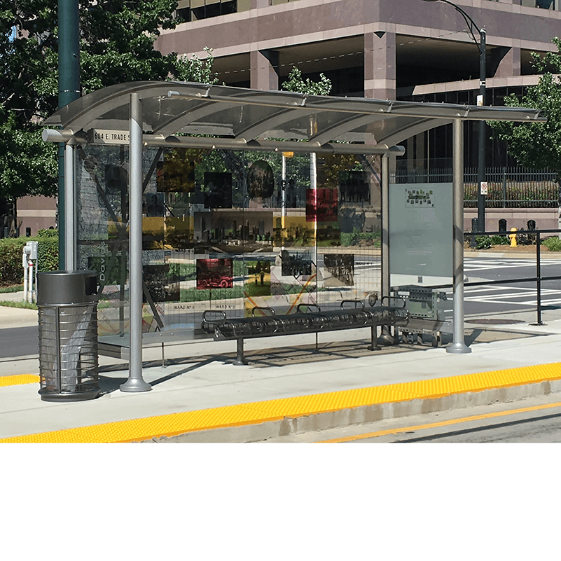 Left side view of a bus shelter displaying a custom design glass back wall, bench seating and a trash receptacle just outside the shelter
