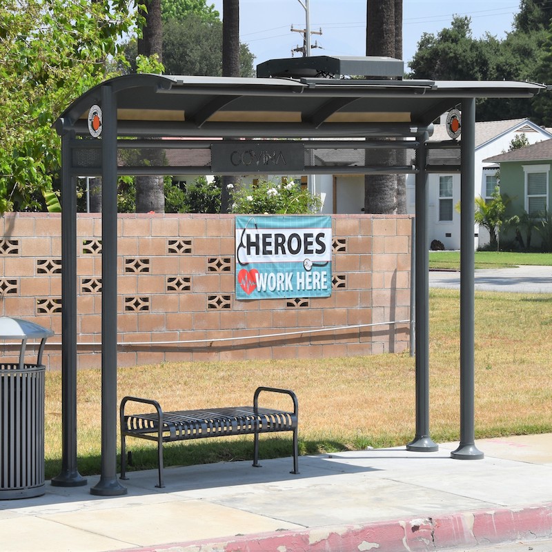A bus shelter sits in Covina with a bench for passengers on the right side along with a trash receptacle. Behind the shelter is a brick wall with "Heroes work Here" banner