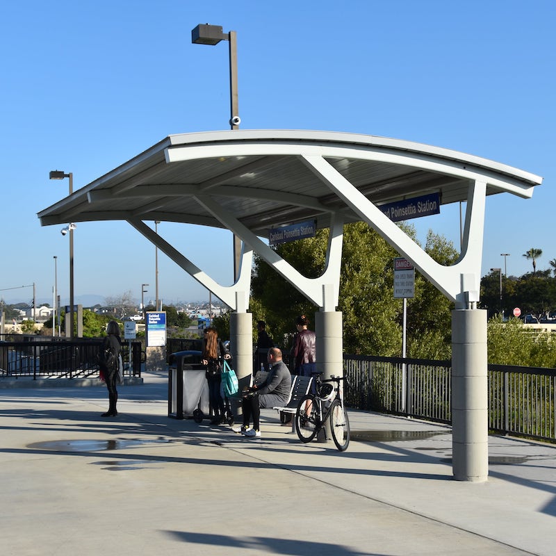 Passengers await the next bus as they sit on the bus shelter's bench, rest their bike and stand in the shelter's shade.