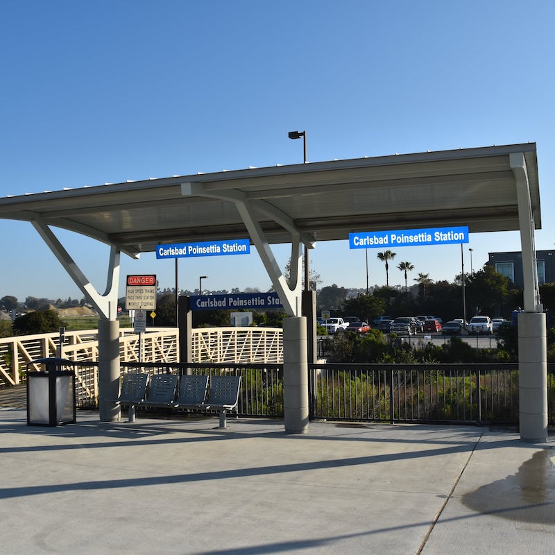 A bus shelter at the Carlsbad Poinsettia Station is seen from the front with seating for four and a trash receptacle.