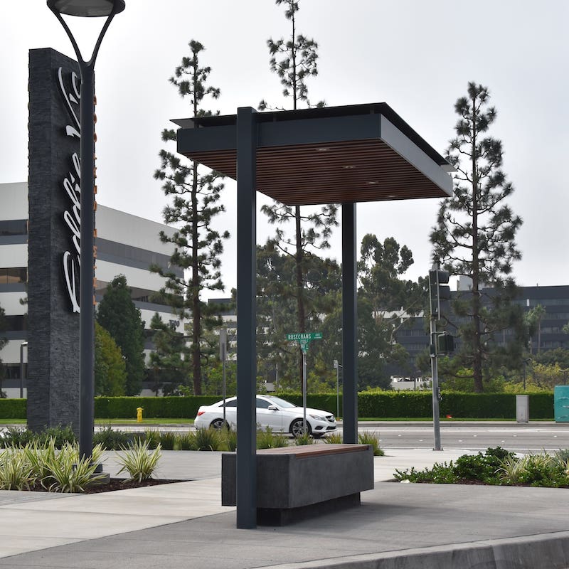 A bus shelter is seen from the left side with a modern design including wood slats on the top of the bench as well as the inside of the roof.