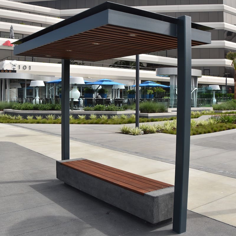 A bus shelter seen from the right side includes a modern design with wood slats on the top of the bench as well as on the underside of the roof.