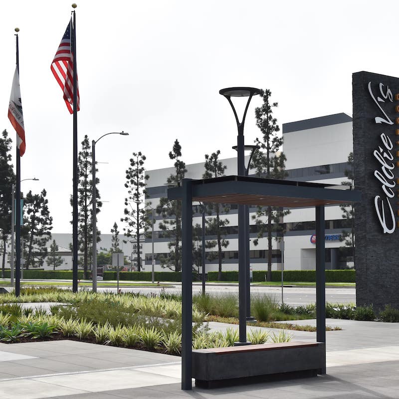 A bus shelter is seen from the front left with a modern design featuring wood slat design on the bench top as well as inside the top of the roof.