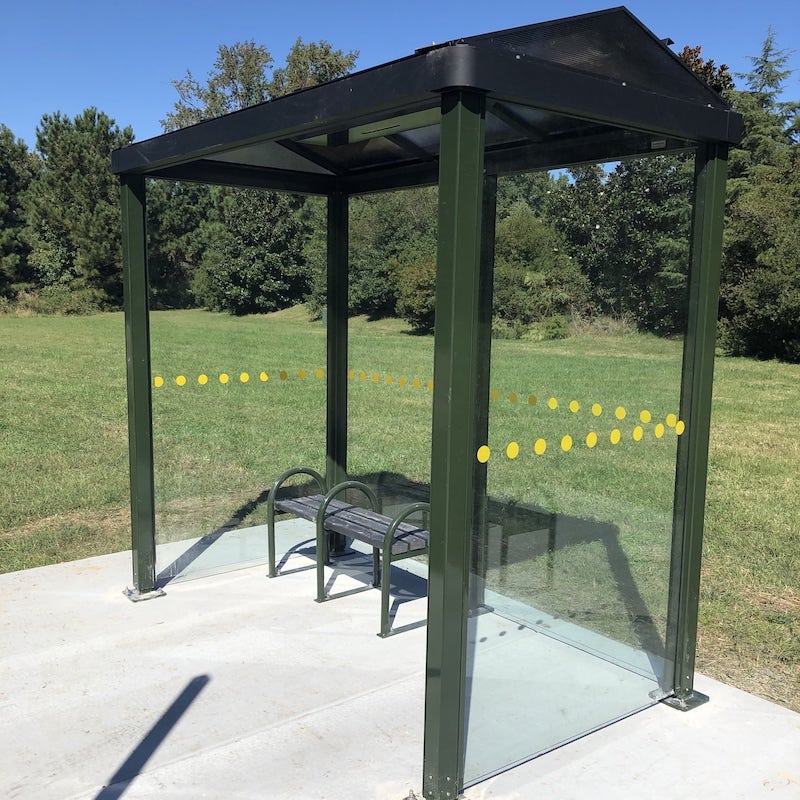 A bus shelter seen from the right front with bench seating for two and a yellow dot design around the middle of the glass panels.