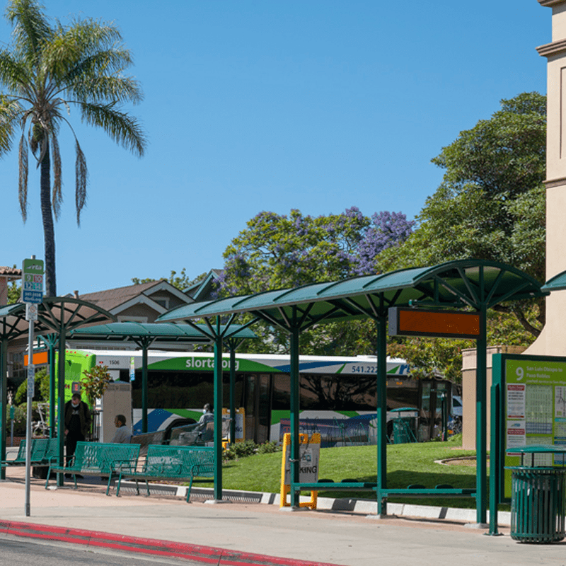 Two bus shelters seen from the front right with three different types of bench seating and a rider information display on the right before the trash receptacle.