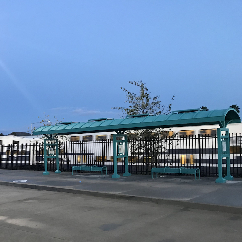 Transit shelters next to each other outside the fence from a passenger train with bench seating under each.
