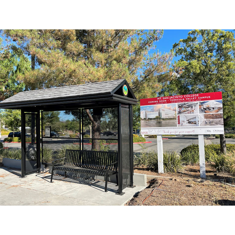 A left side view of a black bus shelter with a grey tile roof, bench seating and a coming soon sign for a new college campus on the left of the shelter