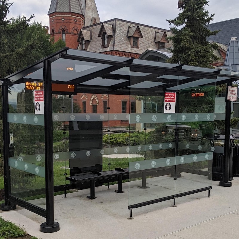A signa orion bus shelter is seen from the front left with glass siding and bench seating inside.