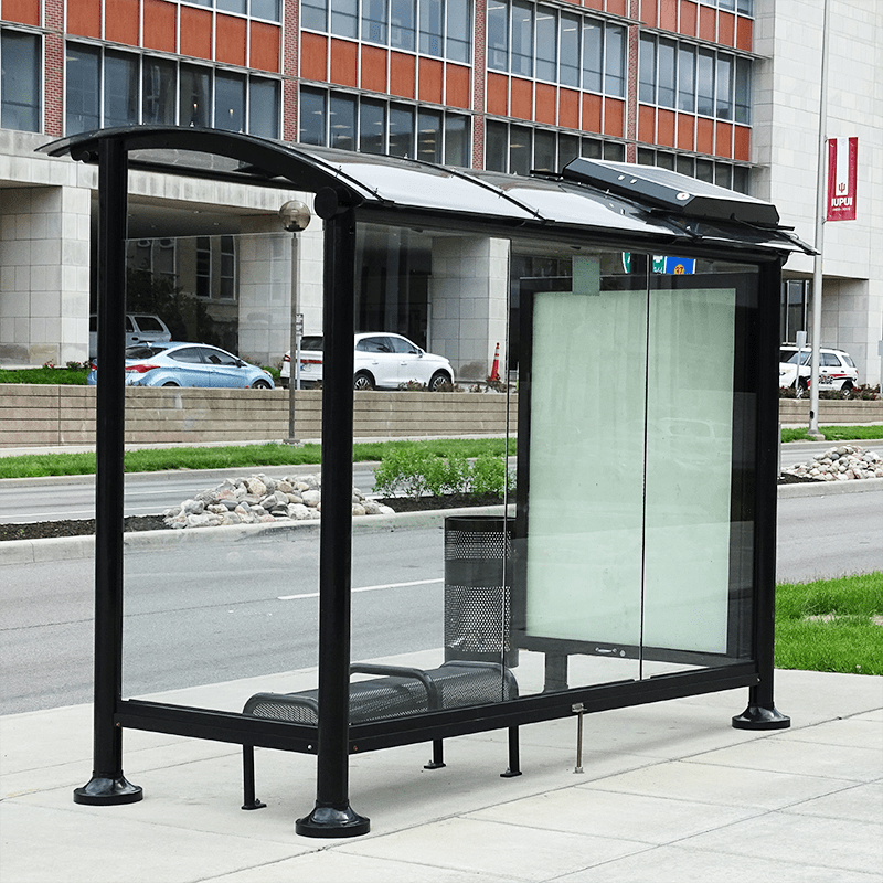 A bus shelter seen from the back left with glass siding showing the inside of the shelter with bench seating and an advertising display on the right side panel.