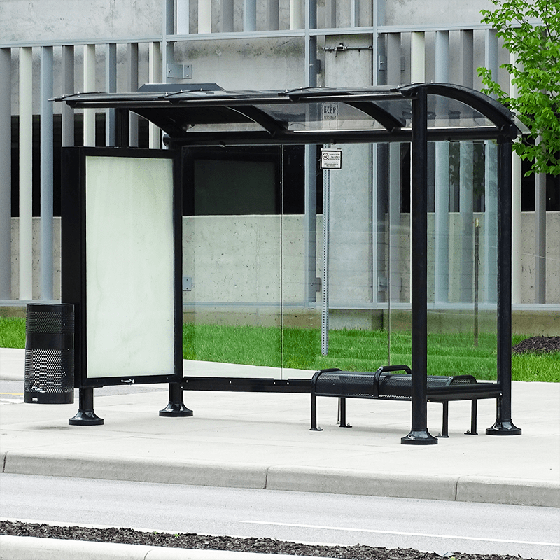 a bus shelter seen from the front right with glass siding, a bench for two inside the shelter, an advertising display on the left panel and a trash receptacle just outside the shelter.