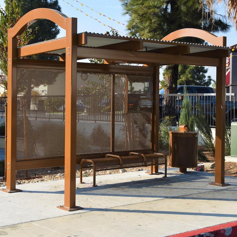 A custom designed mission inspired bus shelter in brown with bench seating for three and a trash receptacle on the outside right.