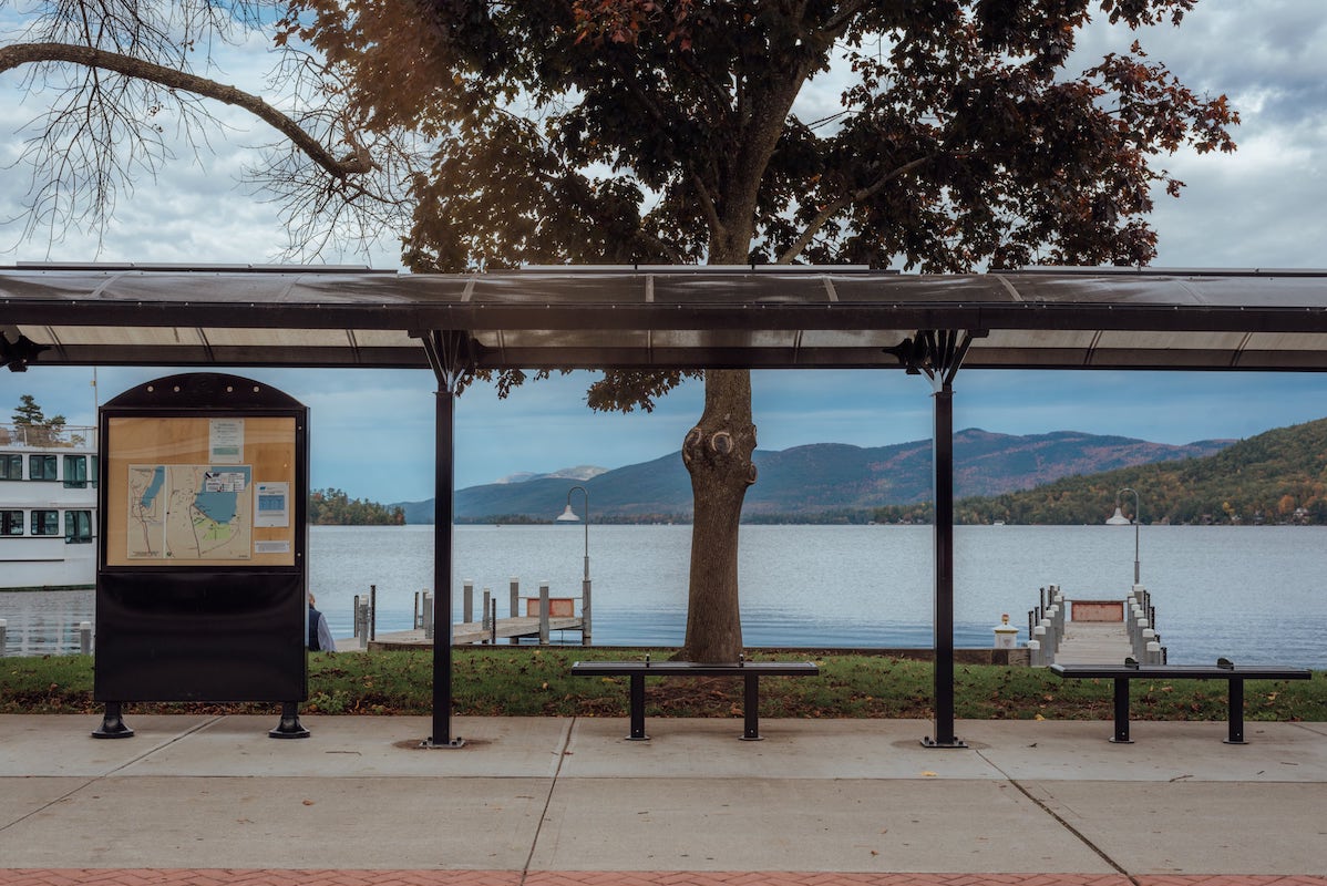 A bus shelter is seen from the front with a lake as the backdrop with a rider information kiosk and two benches for rider seating.
