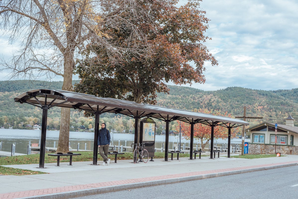 A passenger is seen leaning on one of the posts under a large bus shelter with five benches and a rider information display in the middle.