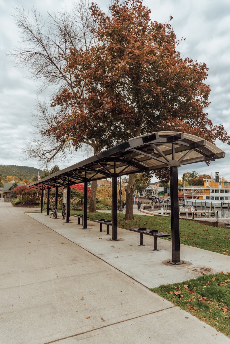 A bus shelter is seen from the front right side with bench seating, a information display kiosk in the middle and a dock and lake in the background