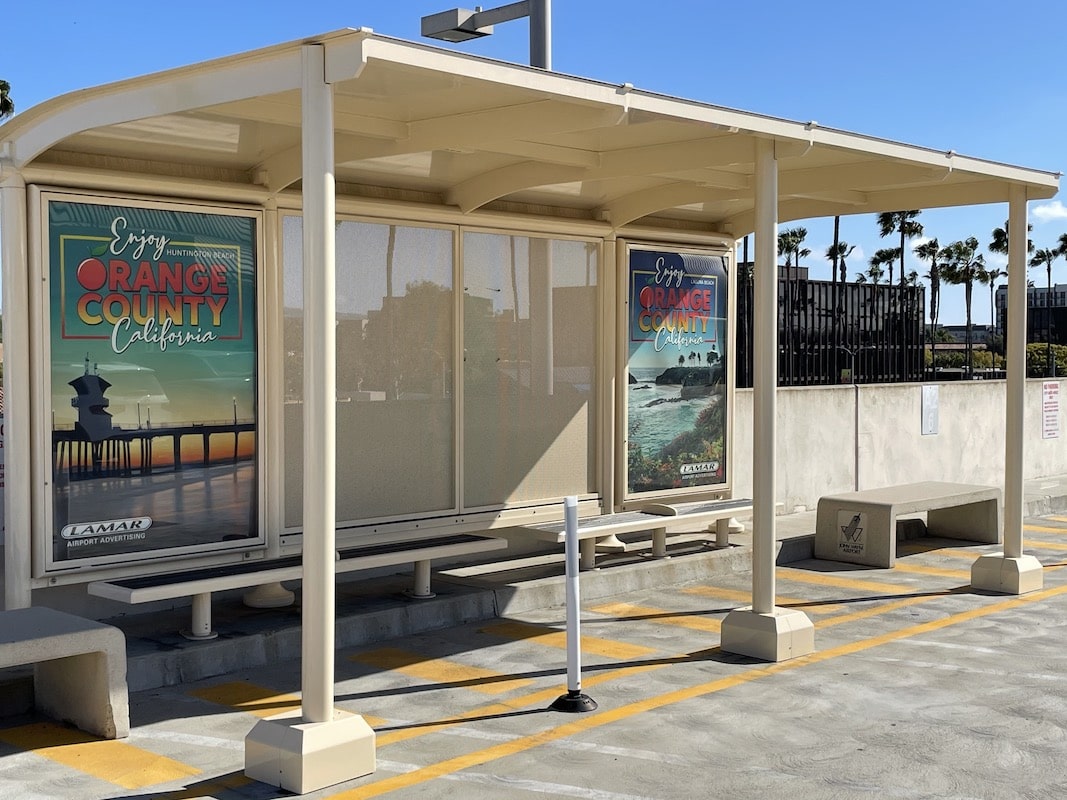 A bus shelter is seen from the left side front with two advertising displays on the back side panels and two benches under the shelter and two outside.