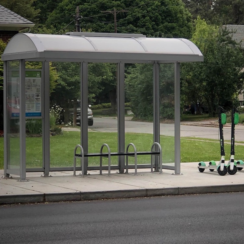 A bus shelter is seen from the left front with bench seating inside, a rider information display inside and lime street scooters on the outside.