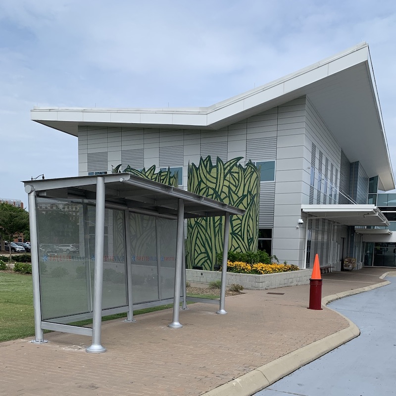 A bus shelter is seen from the front left outside a two story building.