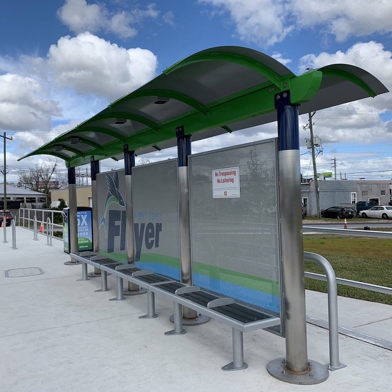 A front right side view of a bus shelter with bench seating spanning the shelter and a trash receptacle on the outside left.