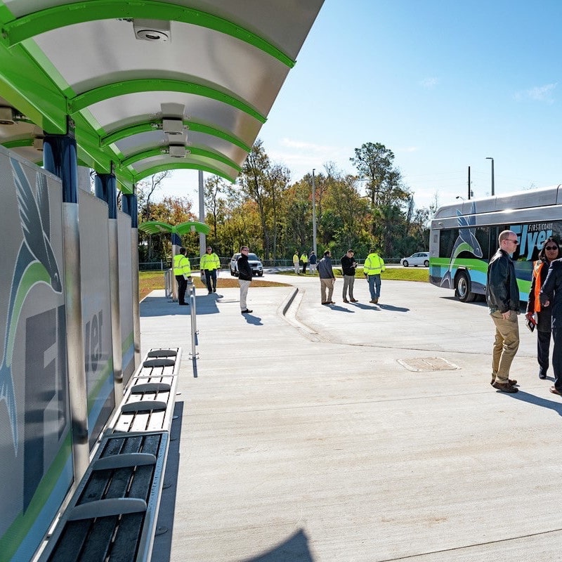 A bus shelter is seen from the left side with passengers and workers standing along the sidewalk and under the shelters.