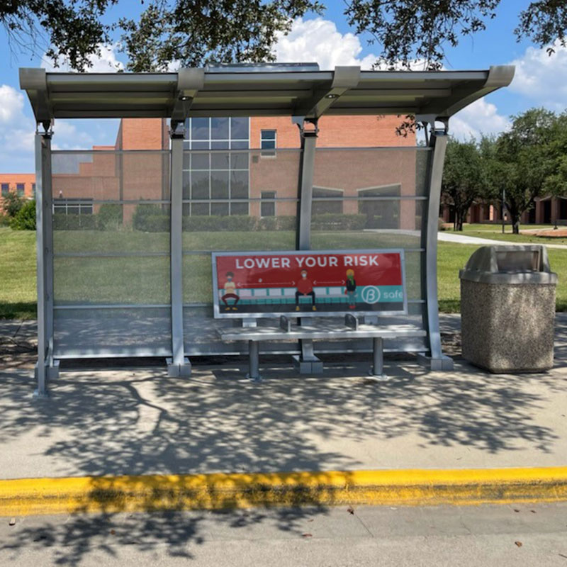 A signa crescent bus shelter seen from the front with an arched roof and structure design, bench seating for three with an advertising back rest and a trash receptacle to the right.