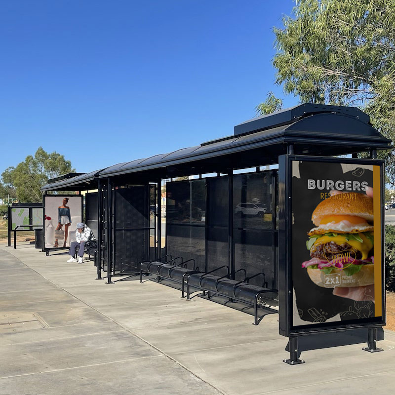 Two bus shelters are seen from the front right side with a rider waiting a ride on the left side shelter next to an advertising display of a woman and the shelter to the right with a right panel advertising display with a burger pictured.