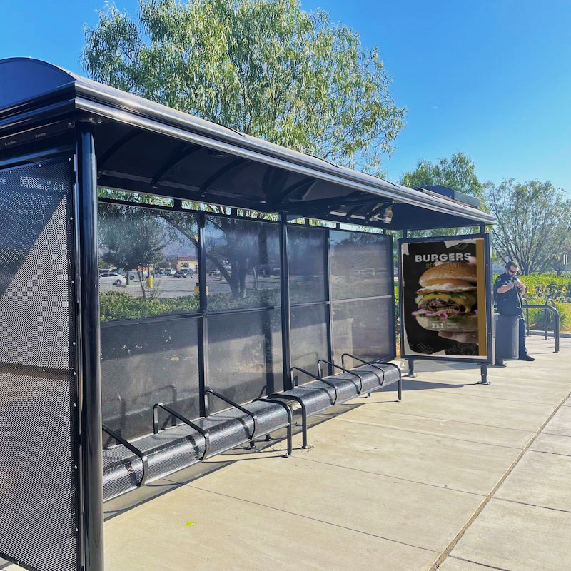 A sierra bus shelter is seen from the front left side with bench seating for eight and an advertising display on the right side panel.