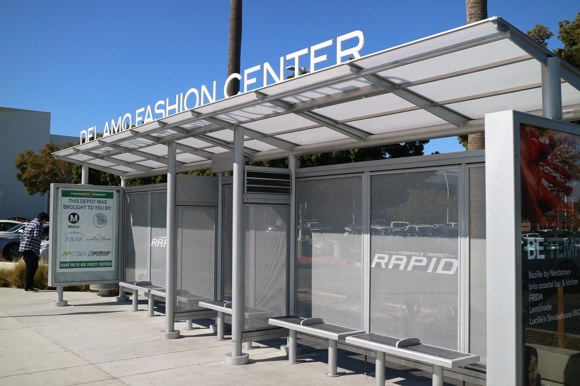 A bus shelter for Del Amo Fashion Center is seen from the front left with three benches and a double sided advertising display on the left side panel.