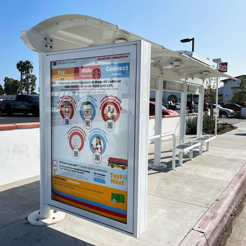 A siga custom bus shelter is seen from the front left side with an advertising display on the left and bench seating on the right under the shelter.