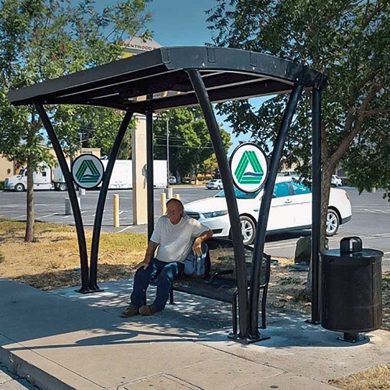A signa custom bus shelter with a rider seated on the bench located under the shelter and a trash receptacle on the right side.