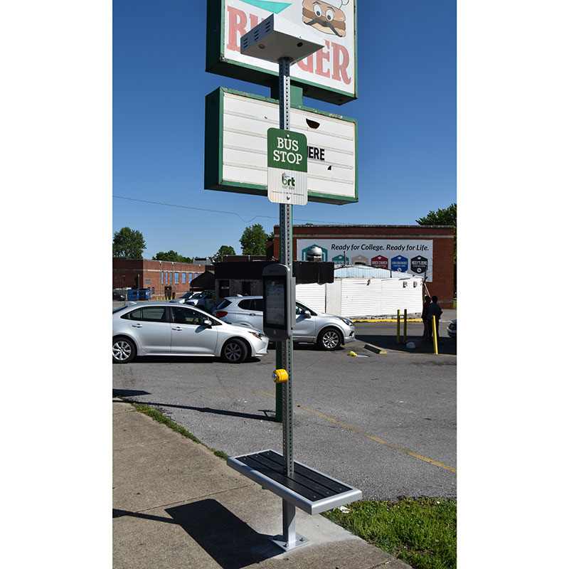 A bus stop post is seen with bench seating attached, rider information displayed on the post and a light on top.