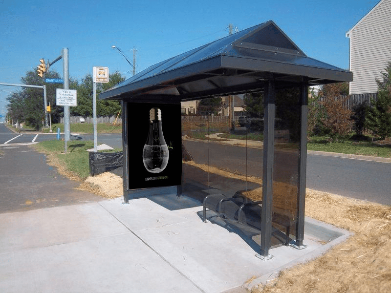 A bus shelter is seen alongside a road with an advertising display on the left pabel and bench seating for three.