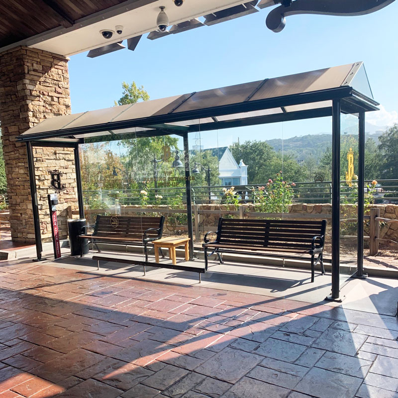 A bus shelter is seen on a brick platform with glass siding, two benches and a side table in between.