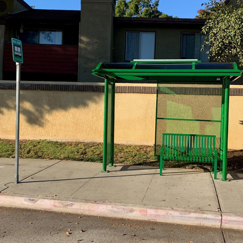 A green bus shelter is seen from the front with a bus stop sign on the left alongside the road and the shelter on the right with bench seating for two.