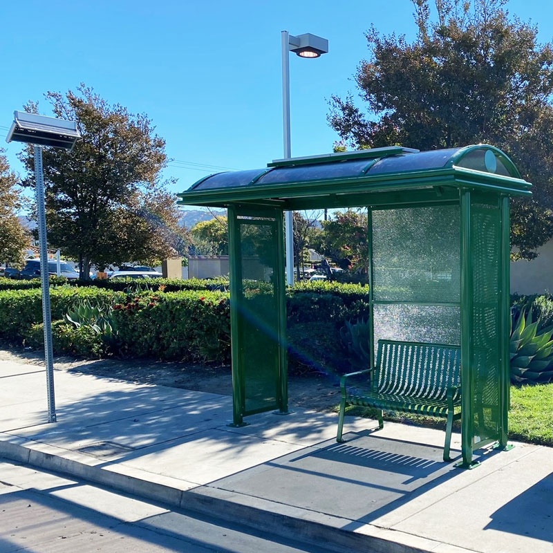 A green bus shelter seen from the front right with a bus stop sign on the left and bench seating for two under the shelter.