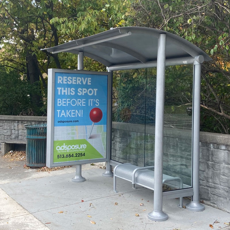 A siga bus shelter is seen from the front right with a trash receptacle on the outside left, an advertising display on the left panel and bench seating under the shelter.