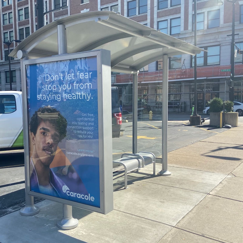 A bus shelter is seen from the left side with a left panel advertising display and bench seating in the shelter for two.