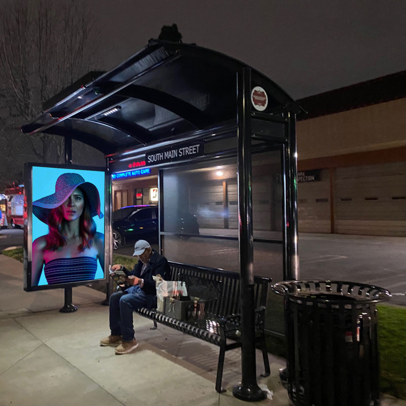 A signa sunset bus shelter seen at night with a lit up bench with a rider seated and an advertisement on the left side panel lit to be seen at night.