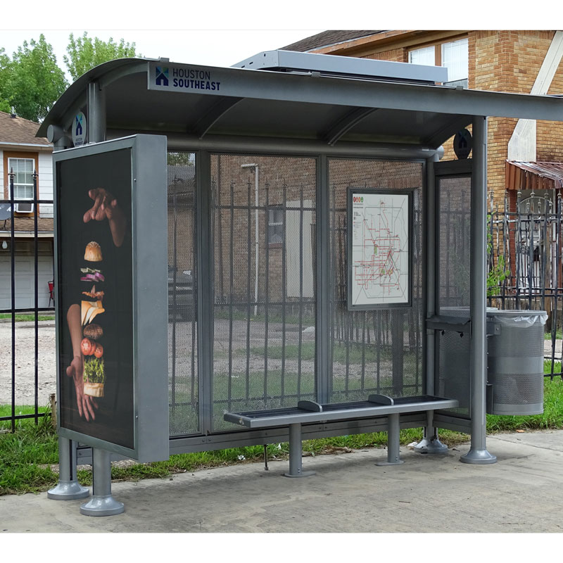 A siga sunset bus shelter is seen with a double sided advertising display on the left, bench seating inside the shelter, rider information on the inside right back panel and a trash receptacle attached to the outside right.