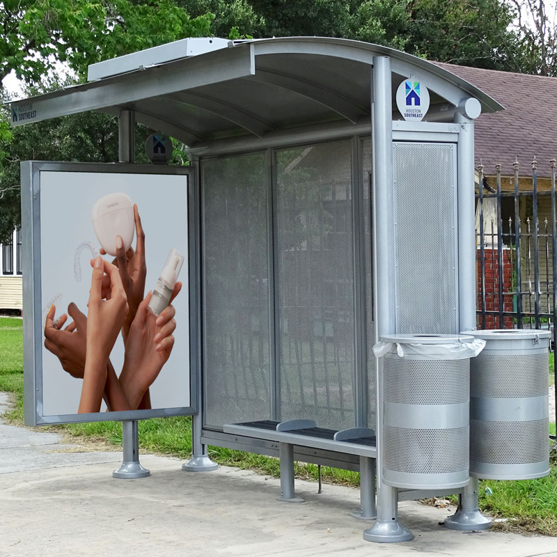 A siga sunset bus shelter is seen from the front right with an advertising display on the left side, bench seating inside the shelter and two trash receptacles on the outside right of the shelter.