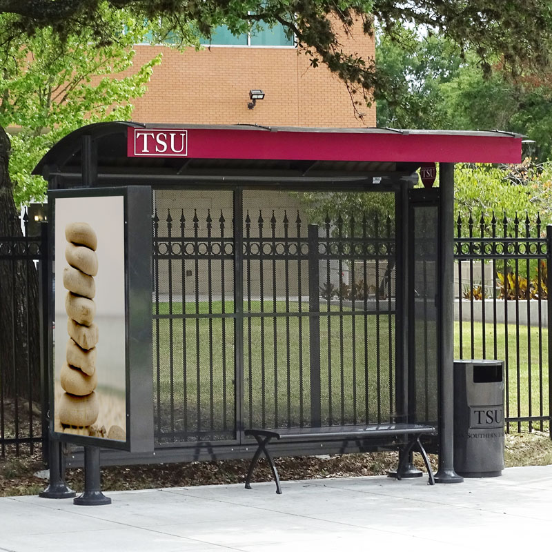 A TSU bus shelter is seen with an advertising display on the left side panel, bench seating under the shelter and a trash receptacle on the outside right.
