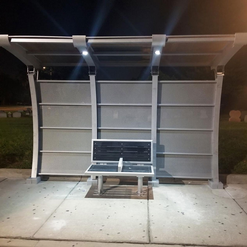 A signa crescent bus shelter seen from the front with overhead lights on from the shelter's roof lighting up a bench for two.