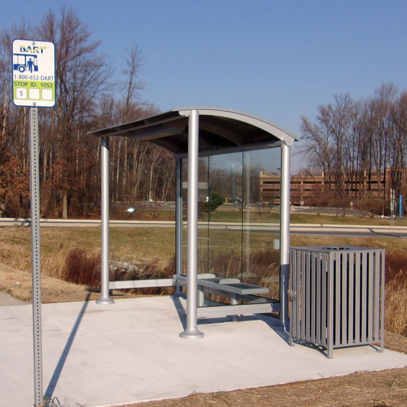 A signa sunset bus shelter is seen from the front right with glass panel siding, bench seating for three and a trash receptacle on the outside right.