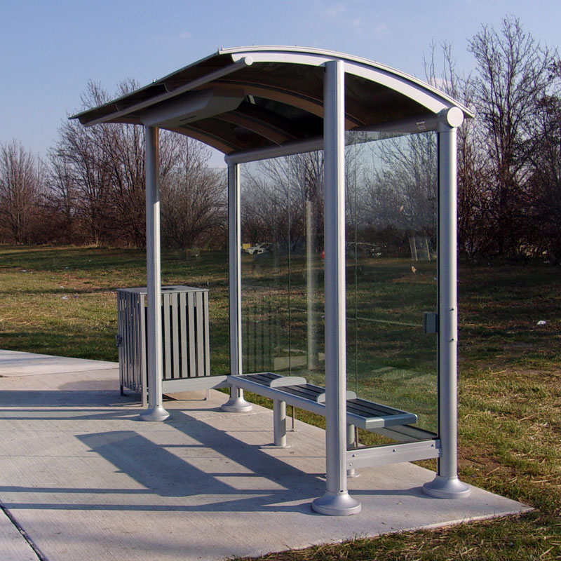 A signa sunset bus shelter is seen from the front right with glass panel siding, bench seating for three and a trash receptacle on the outside left.