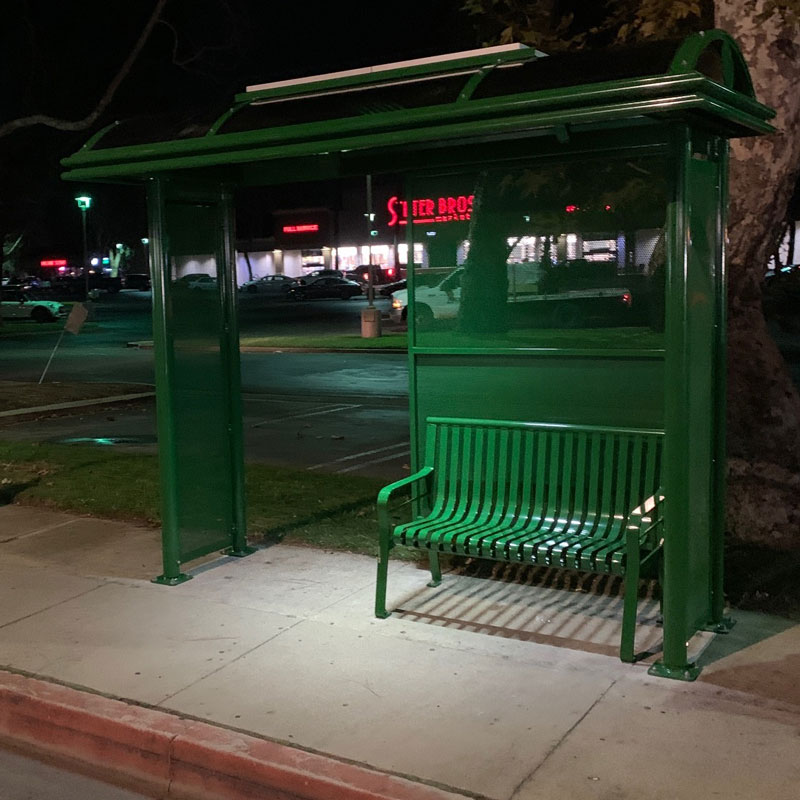 A green bus shelter is seen from the front right at night with shelter lighting and bench seating for two.