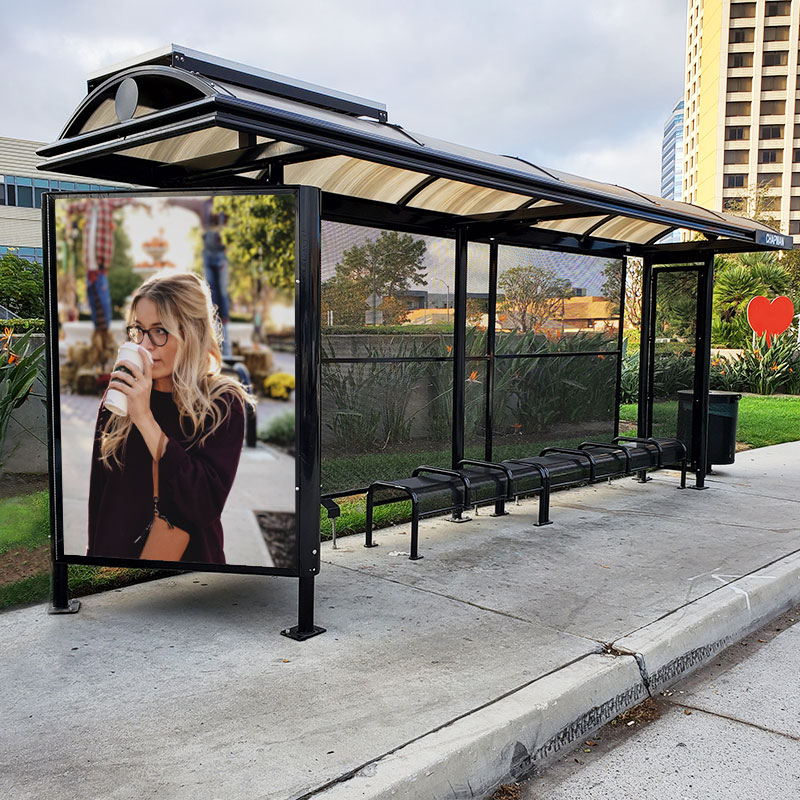 A bus shelter is seen from the front left side with ample bench seating, a left panel advertising display and a right side trash receptacle.