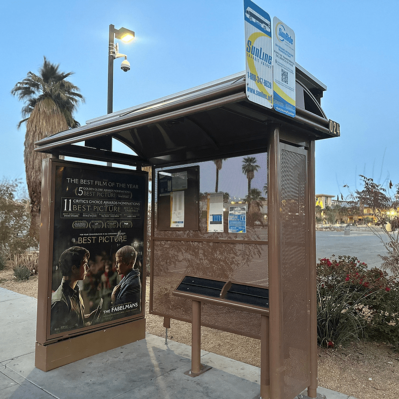 A sierra bus shelter is seen from the front right with a left side panel advertising display, rider information posted inside and a standing bench.